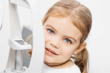 A pediatric optometrist examines the eyes of a little girl using modern equipment. The happy girl...