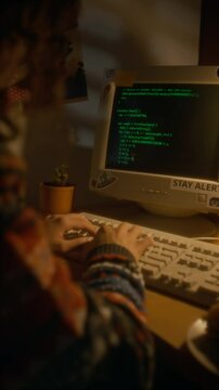 Vertical over shoulder shot of millennial developer in sweater typing programming code on old retro computer at desk in evening, working at home in 90s