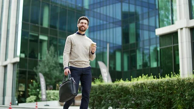 Businessman in casual attire walks outside modern glass office building holding coffee cup and briefcase symbolizing success and morning routine. Urban setting reflects professional atmosphere