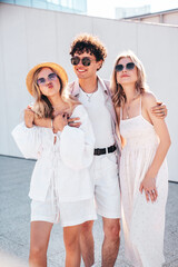 Young three stylish friends posing in the street. Fashion man and two cute female dressed in casual summer clothes. Smiling models having fun. Cheerful women and guy outdoors, in sunny day, sunglasses