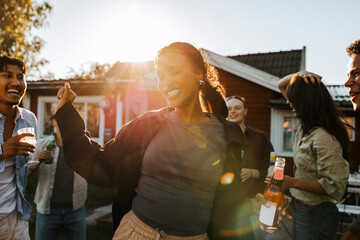 Carefree young women holding beer bottle while dancing with friends in back yard on sunny day at social gathering