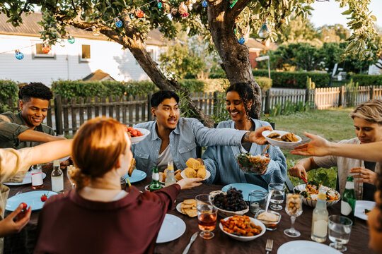 Male and female friends passing food to each other while having dinner in back yard at social gathering - Powered by Adobe