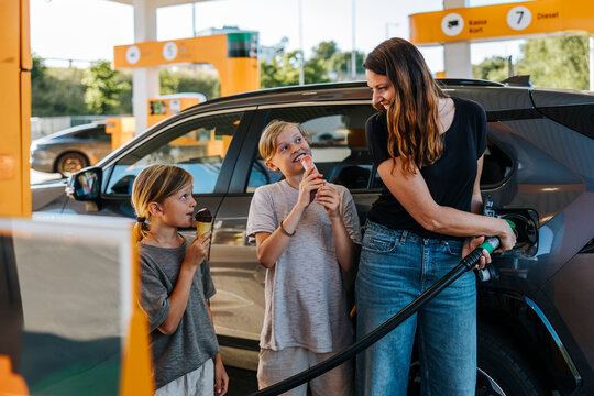 Smiling woman refueling car and standing with sons eating snacks at gas station