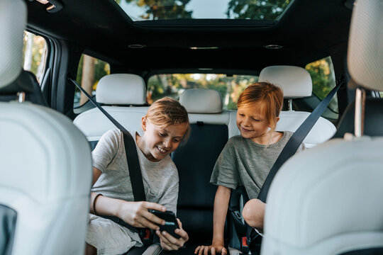 Happy male siblings spending leisure time watching smart phone while sitting in car during road trip