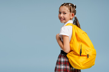 Schoolgirl with yellow backpack smiling over shoulder on blue background