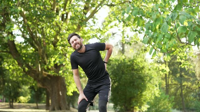 Sportsman suffering from back pain, sports injury, muscle spasm during a morning run in the urban city park. Adult bearded runner having back flank ache and problem after exercise outside in nature