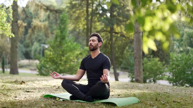 Adult man meditating outdoors in peaceful park environment. Seated on yoga mat amidst trees, focusing on calmness and mindfulness. Concentrated male practices meditation barefoot with his eyes closed