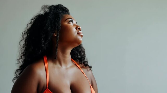 Confident young woman with natural curly hair looking upwards in thoughtful pose, wearing an orange top, showcasing body positivity and self-confidence in a minimalist setting.