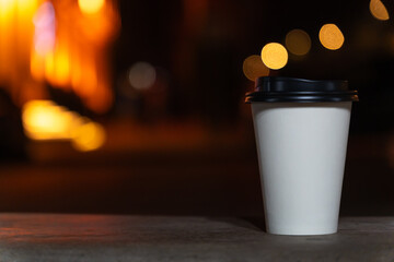 Close-up of a disposable white paper cup with coffee or tea stands on a bench against the background of a blurred city at night with lights. Coffee to go