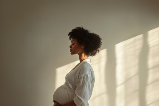 Side view of a pregnant woman standing in sunlight and holding her belly