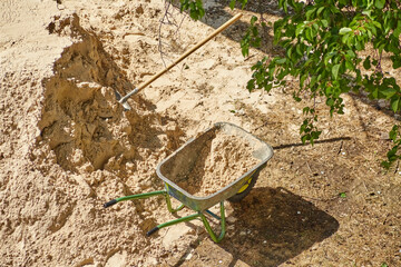 A shovel in a pile of sand and a wheelbarrow with sand under a tree, construction work on a plot,...