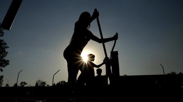 The silhouette of a sporty, pumped-up girl on an orbitrek on an outdoor simulator in the park, the summer setting sun. A sporty woman with a toned body trains on an outdoor sports field. Slow motion.