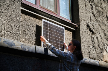 Woman holding photovoltaic solar panel in front of historical building, wearing plaid shirt and...
