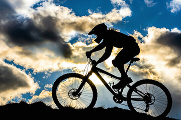 Obraz premium silhouette of cyclist against dramatic cloudy sky, sun peeking through