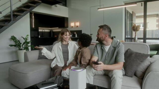 Full shot of young Caucasian couple sitting on sofa in living room with adoptive African American boy, teaching to use smart home system, saying command into interactive speaker and lights turning on