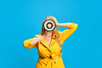 Fototapeta premium Blonde woman in yellow dress holding megaphone against blue studio background. Retail announcement, flash sales, Black Friday promo. Concept of sales, shopping, news, announcement, advertisement