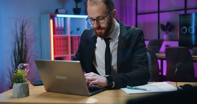 Businessman in formal wear and in glasses working on laptop computer at late time in office sitting at his workplace