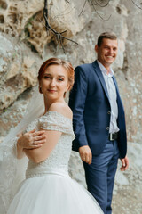 A bride and groom are posing for a picture in front of a rock wall. The bride is wearing a white dress and the groom is wearing a blue suit. They both look happy and are smiling for the camera