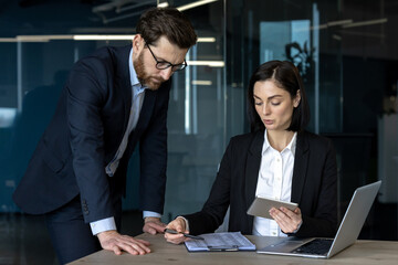 Business professionals engaged in workplace discussion over project details. Woman uses tablet for reference. Man leans over table, emphasizing teamwork. Office environment highlights technology