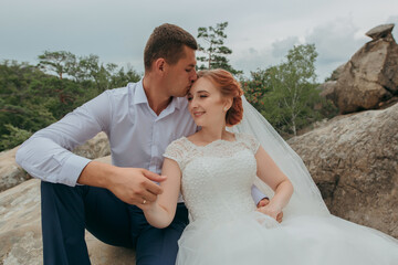 A bride and groom are sitting on a rock, kissing each other. The bride is wearing a white dress and the groom is wearing a white shirt