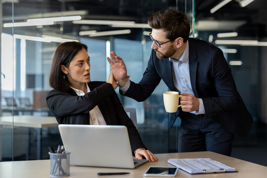 Businesswoman sitting at desk uses strong body language to halt unwanted advance from male colleague with coffee cup, illustrating workplace dynamics, conflict, and harassment prevention.