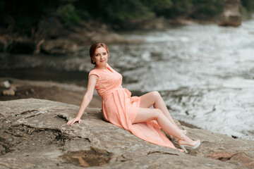 A woman in a pink dress is sitting on a rock by the water. The scene is serene and peaceful, with the woman posing for a photo
