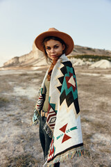 Lonely woman in hat and scarf standing in desert with blanket draped around shoulders