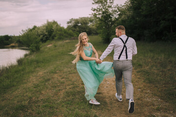 A young couple is running through a field, with the woman wearing a green dress. The man is wearing a white shirt and black suspenders. Scene is playful and romantic