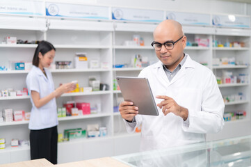 Asian professional male pharmacist using digital tablet computer checking drug list Pharmacist checking stock in pharmacy for health medicine in pharmacy with female pharmacist in the background
