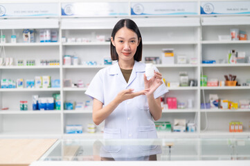 Asian professional female pharmacist standing with arms crossed looking at camera charming smile pointing hand to pill bottle health care products in pharmacies Close-up and wide-angle shots