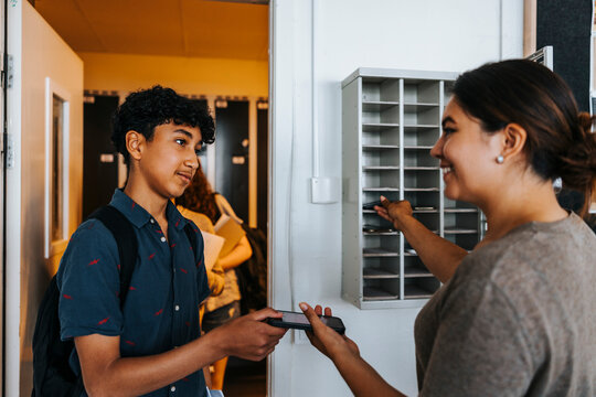 Smiling female teacher collecting smart phone from teenage boy standing near doorway of classroom at school