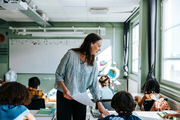 Smiling female professor distributing test papers to pupils sitting in classroom at school