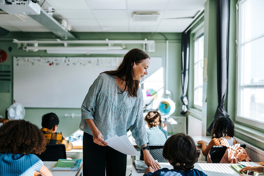 Smiling female professor distributing test papers to pupils sitting in classroom at school
