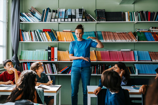 Female teacher gesturing and explaining group of elementary students while standing in front of bookshelf at library