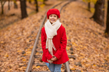 Caucasian girl in a red coat and beret walks along the railway tracks in the park in autumn.