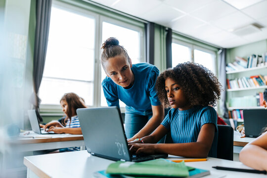 Female teacher assisting curly hair girl studying on laptop while sitting near desk in classroom at elementary school