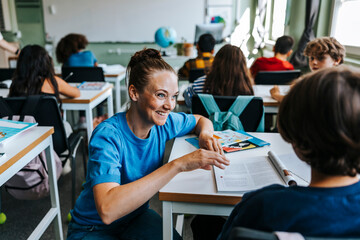 Smiling female teacher assisting student while crouching near desk in classroom at school