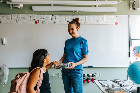 Smiling female teacher collecting smart phone of schoolgirl while standing in classroom at elementary school