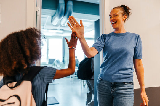 Cheerful female teacher giving high-five to female pupil while standing near doorway of classroom in school
