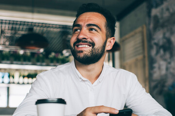 Positive man in casual formal shirt sitting at cafeteria table with modern cellphone looking away and smiling from funny information on financial website, bearded male manager feeling good indoors