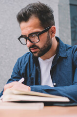 Concentrated male student in classic spectacles for vision correction sitting at street cafeteria and learning preparing to university exams, bearded man making memo notes in knowledge textbook