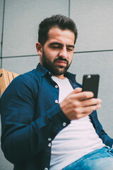 Millennial hipster guy in casual outfit sitting outdoors and spending time for coffee break reading received notification on cellphone gadget, bearded man updating profile during networking via mobile