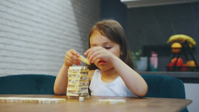 Tabletop game, young child joyfully engages with a wooden block tower game in cozy atmosphere of their home kitchen, showcasing their independence and creativity as they build dreams, happy memories