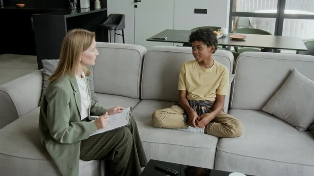 Full shot of young Caucasian female social worker or psychologist with clipboard interviewing 11-year-old black boy, discussing life in new foster home and relationship with family members