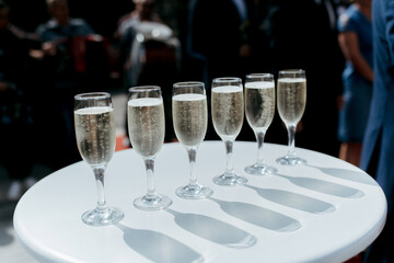 A table with six champagne glasses on it. The glasses are all filled with champagne and are lined up in a row. The table is white and the glasses are clear. The scene is elegant and sophisticated