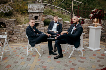 Three men in suits are sitting on chairs in a courtyard. One of the men is wearing a tie and a pink...