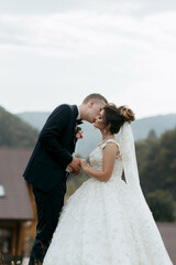 A bride and groom are kissing each other on the cheek. The bride is wearing a white dress and the groom is wearing a suit. The scene is set in a mountainous area