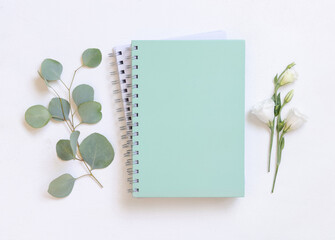Blank spiral notebook on a white table near green eucalyptus leaves and flowers, school mockup