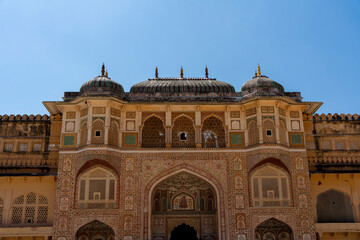 the grand Ganesh Pol at Amber Fort in Jaipur, India. The intricately decorated gateway showcases vibrant floral motifs, delicate latticework, and domed pavilions against a bright blue sky.