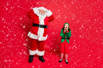 Full body photo of astonished santa claus with his granddaughter in green elf costume scream omg see noel incredible miracle wearing belt eyewear eyeglasses isolated over red background